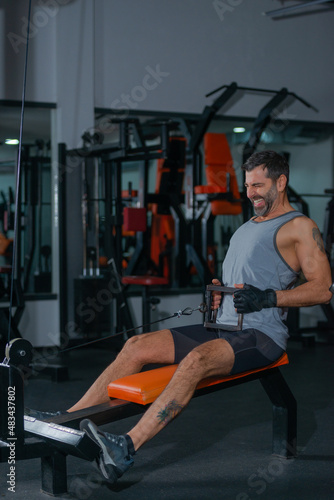 Fitness training in sport gym of active male with beard sitting and pulling heavy weight on exercise equipment