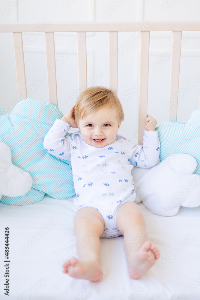 a cheerful blonde baby boy in a crib at home with blue and white cotton bedding smiles in the morning, the concept of children's goods and accessories a cheerful blonde baby boy in a crib at home with blue and white cotton bedding smiles in the morning, the concept of children's goods and accessories