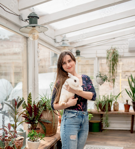 Young woman holding a white rabbit in her arms while standing in a home flower greenhouse