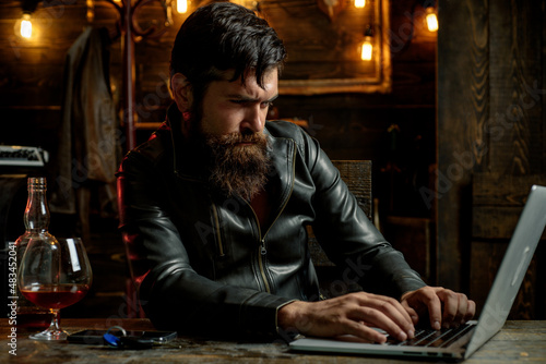 Bearded man drink cognac. Drunk attractive handsome sad pensive man in black jacket, business man drinking whiskey in front of laptop at loft work place.