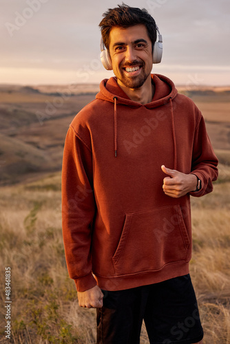 Portrait of smiling sportsman in headphones in summer filed in the morning at sunrise, bearded guy in red casual hoodie having rest after jogging, running outdoors, relaxing, listening to music.