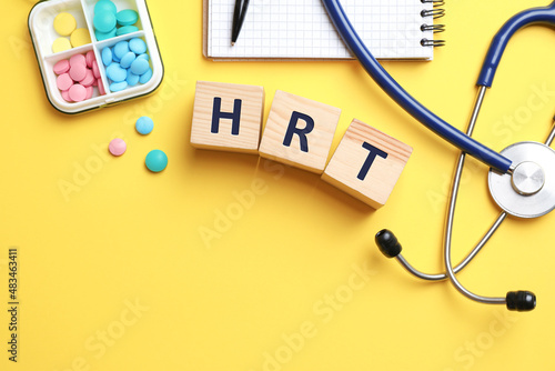 Wooden cubes with abbreviation HRT, pills, stethoscope and notebook on yellow background, flat lay. Hormone Replacement Therapy photo