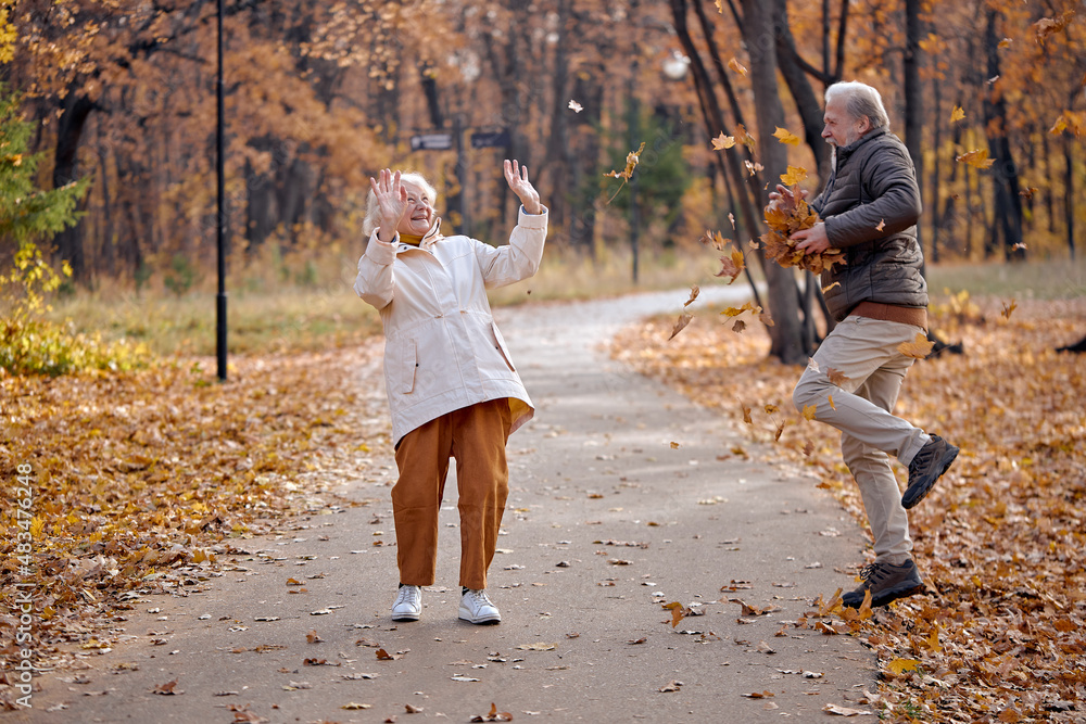 Fototapeta premium joyful senior couple having fun together in park while walking, man throwing leaves up. Caucasian european man and woman in coats laughing, enjoy spending time together. lifestyle, human emotions