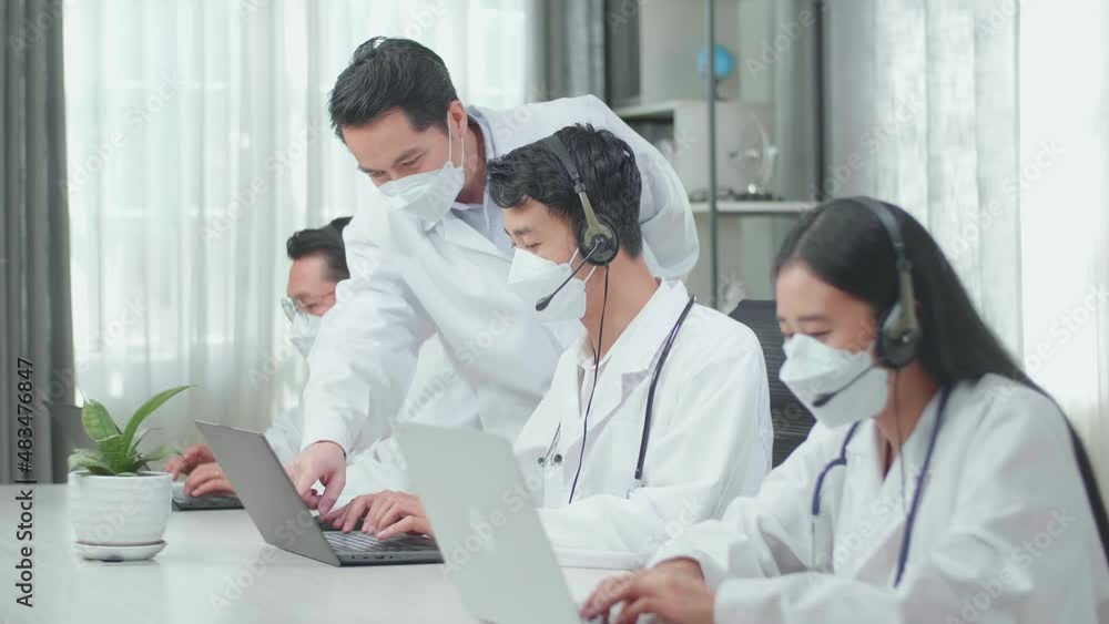 A Man Coming To Help One Man Of Three Asian Doctors In Headsets And Masks Using Computer For Working As Call Center Agent While His Colleagues Are Speaking And Typing During A Call At The Office.
A Man Coming To Help One Man Of Three Asian Doctors In Headsets And Masks Using Computer For Working As Call Center Agent While His Colleagues Are Speaking And Typing During A Call At The Office.