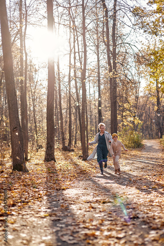 Positive mom and child boy running, having fun in forest or park, laughing together. Caucasian family kid boy and mother in coats enjoy the sunny warm weather at autumn season, leaf fall © Roman