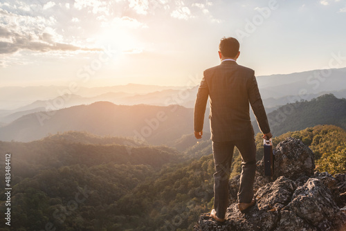 Successful businessman holding his business attache on top rock mountain sunset background, competition and leadership concept.
