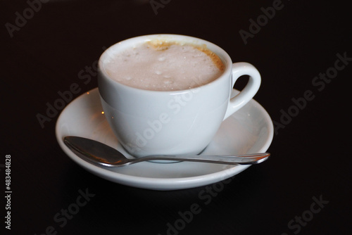 a natural cappuccino in a white cup on a saucer with a small spoon stands on a black table. side view