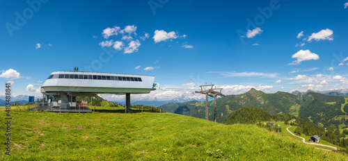 Beautiful view of alpine mountain scenery, blooming green meadows, snowcapped mountain peaks, mountain cable car station. The sunny day with blue sky in summer.