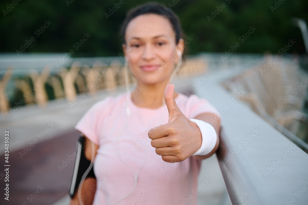 Soft focus on a hand showing thumb up of a beautiful out of focus female athlete in pink t-shirt, smiling standing on a treadmill on the city bridge while working out outdoor Soft focus on a hand showing thumb up of a beautiful out of focus female athlete in pink t-shirt, smiling standing on a treadmill on the city bridge while working out outdoor