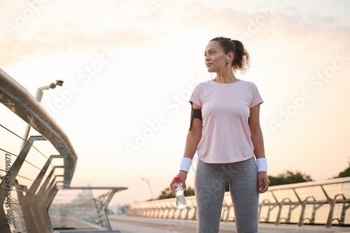 Beautiful athletic woman, female athlete, sportswoman with earphones holds a bottle with water and looks aside standing on a city bridge treadmill early in the morning during her workout at sunrise