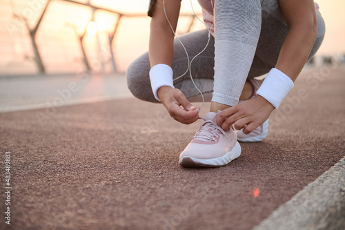 Cropped image of a fitness woman tying shoelaces on sneakers and getting ready for morning running and sports workout outdoors on a city bridge treadmill at sunrise