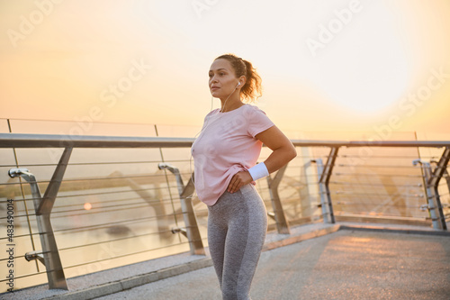 Confident sportswoman, determined female athlete, runner jogger with beautiful healthy body wearing pink t-shirt, gray leggins and white terry wristbands standing on a city bridge treadmill at sunrise