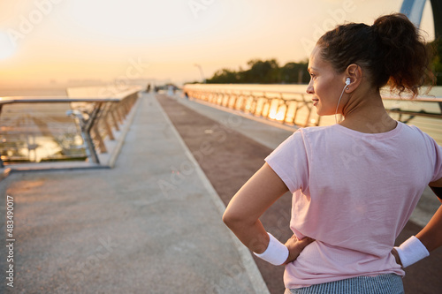 Rear view of a pretty athletic woman, female runner athlete, standing with her hands on waist on the city bridge treadmill and looking away, ready for morning cardio training and jogging at sunrise