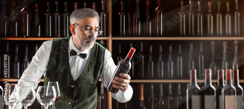 Sommelier with a glass of wine. Examination of wine products. Restaurant staff, expert wine steward among shelves of wine bottles. Stylish middle-aged man with a grey beard photo