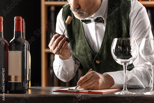 Sommelier with a glass of wine. Examination of wine products. Restaurant staff, expert wine steward among shelves of wine bottles. Stylish middle-aged man with a grey beard photo