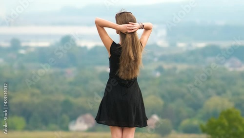 Young pretty woman with long hair in black short dress standing outdoors enjoying view of summer nature