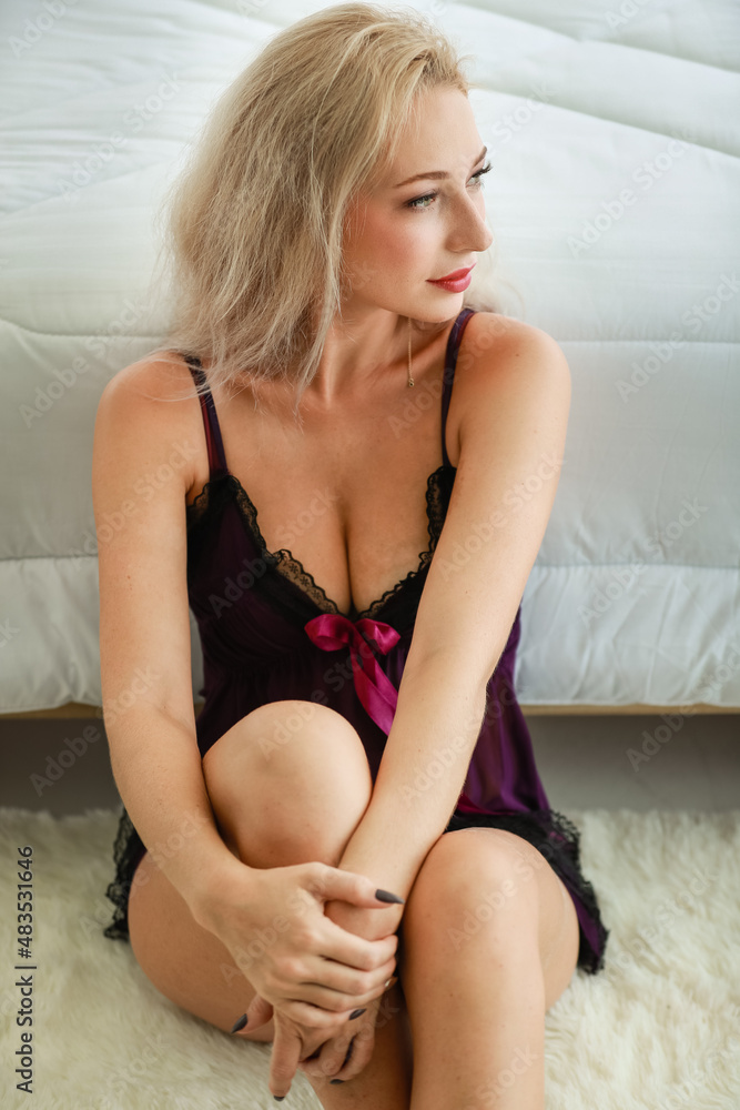 Naklejka premium Portrait studio shot of Caucasian female blonde long hair fashion model in black sexy lingerie underwear sitting on white carpet floor leaning on bed with blanket smiling look at camera in bedroom
