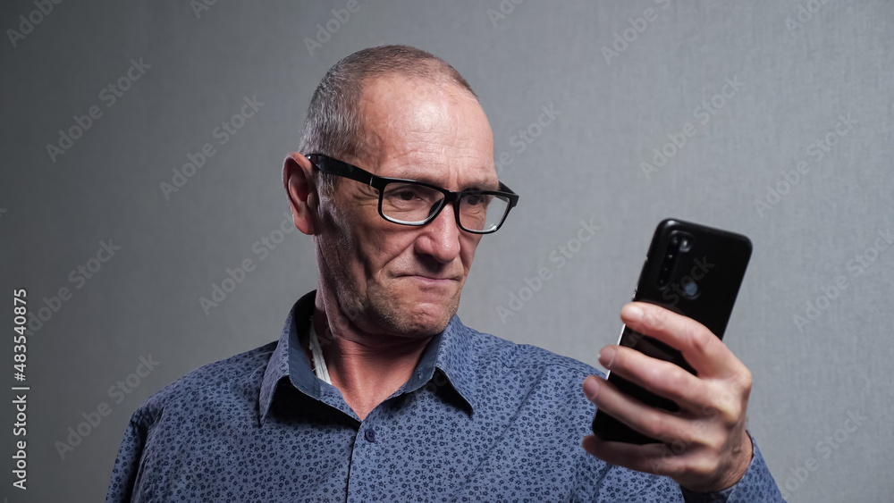 Senior man pensioner with bad eyesight in elegant black-rimmed glasses tries to read message in modern smartphone on grey background closeup. Senior man pensioner with bad eyesight in elegant black-rimmed glasses tries to read message in modern smartphone on grey background closeup.