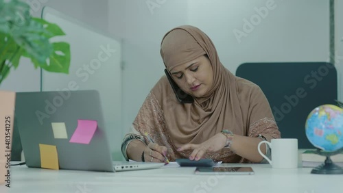 Young indian Muslim businesswoman talking on Mobile Phone Working On Laptop In Modern Office.Asian Muslim businesswo girl talking on file checking and signature. photo