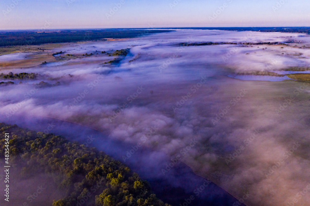 Fototapeta premium Aerial view of a beautiful summer landscape with a fog while dawn. Photo from drone of a foggy landscape in spring. Top view to land while sunrise. Early morning in nature, misty weather.