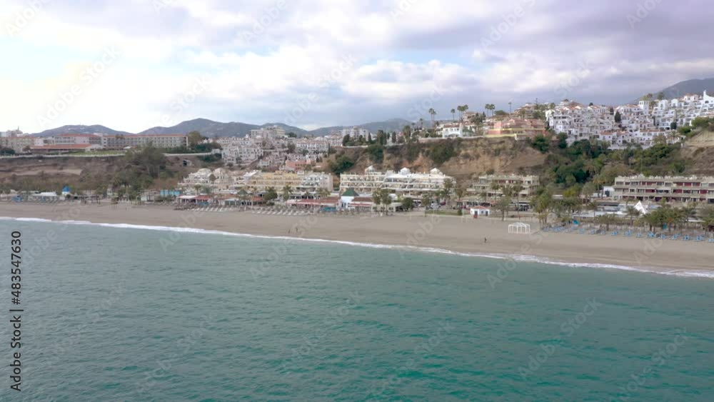 Nerja city, SPAIN. Aerial perspective of Burriana playa in winter. Few people walking on the beach. Drone going forward. Typically white Andalusian houses on hills. Cloudy sky. Bars and restaurants. Nerja city, SPAIN. Aerial perspective of Burriana playa in winter. Few people walking on the beach. Drone going forward. Typically white Andalusian houses on hills. Cloudy sky. Bars and restaurants.
