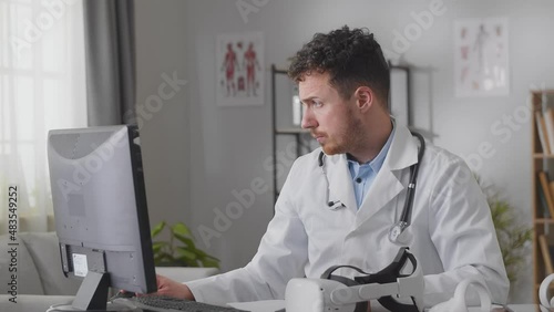 Tired caucasian male doctor in white gown sitting at desk in cabinet and massages his face and neck
