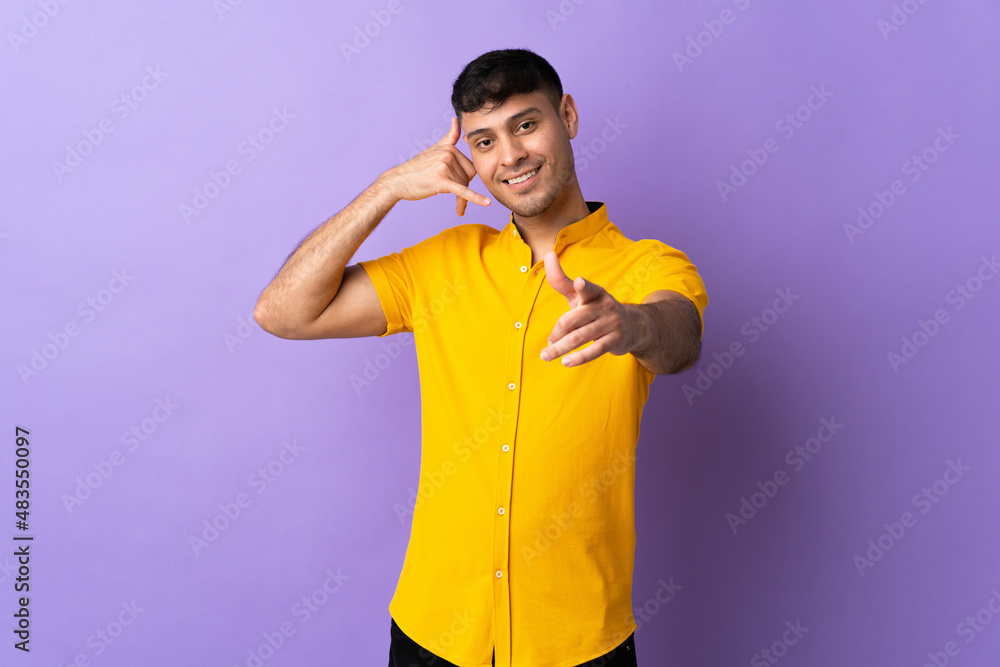 Young Colombian man isolated on purple background making phone gesture and pointing front Young Colombian man isolated on purple background making phone gesture and pointing front