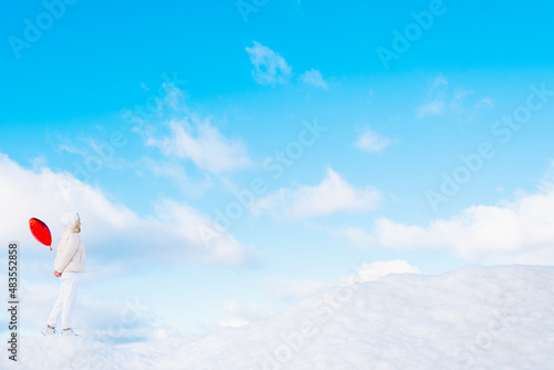 Fototapeta Naklejka Na Ścianę i Meble -  Portrait of young woman in white clothes with red heart balloon walking on snow cover with blue cloudy sky on background. Sky merges with earth near snowdrifts. Side view. Copy space. Minimalism