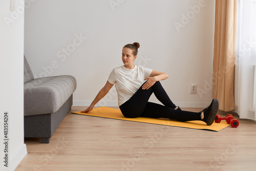 Horizontal shot of beautiful young adult sporty woman practicing yoga at home on yoga mat, posing in living room near sofa and window, turning her body aside, stretching.