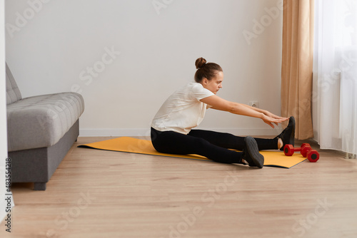 Indoor shot of slim attractive woman wearing white t shirt and black leggins sitting on yoga mat on the floor near sofa and stretching her legs, healthy lifestyle.
