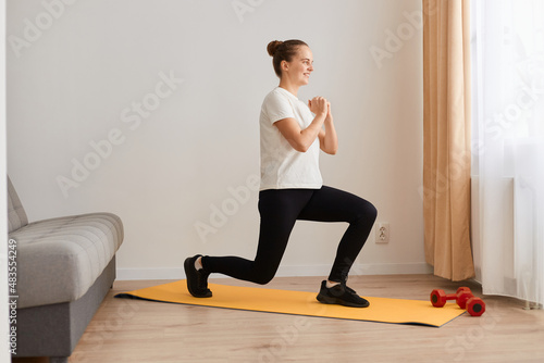 Full length profile portrait of sporty athletic girl doing lungs workout in living room, keeps hand together, looking straight ahead, wearing white t shirt and black leggins.