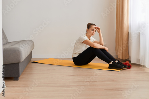 Domestic training. Tired woman exercising at home, taking break, resting and thinking, sitting on yoga mat in living room, wearing white t shirt and black leggins.
