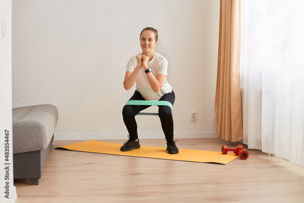 Full length portrait of athletic woman wearing white t shirt and black leggins, doing squats with resistance band on knees, pumping legs and glutes muscles, looking smiling at camera. Full length portrait of athletic woman wearing white t shirt and black leggins, doing squats with resistance band on knees, pumping legs and glutes muscles, looking smiling at camera.