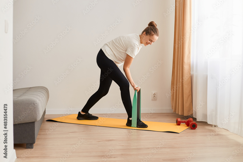 Portrait of sporty woman at home trying to lose weight, standing on rubber stripe training legs, exercising with rubber expander indoor, resistance band for muscle stretching and strength workouts. Portrait of sporty woman at home trying to lose weight, standing on rubber stripe training legs, exercising with rubber expander indoor, resistance band for muscle stretching and strength workouts.