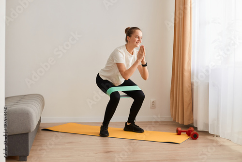 Full length portrait of attractive beautiful charming sporty woman wearing white t shirt and black leggins, doing sit-ups with elastic band, workout at home, fitness and healthy lifestyle. © sementsova321