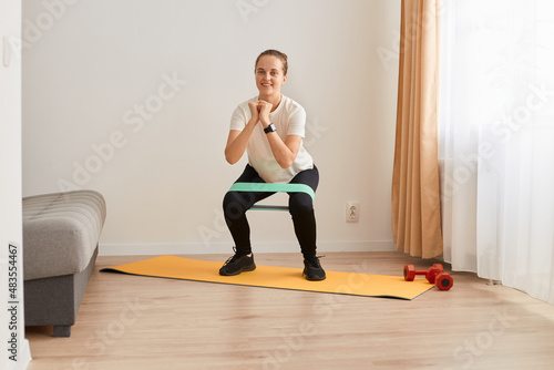 Full length portrait of athletic woman wearing white t shirt and black leggins, doing squats with resistance band on knees, pumping legs and glutes muscles, looking smiling at camera.
