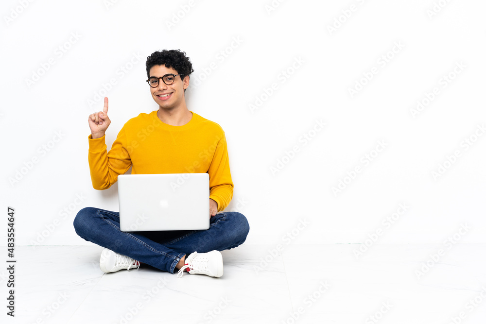 Venezuelan man sitting on the floor with laptop showing and lifting a finger in sign of the best Venezuelan man sitting on the floor with laptop showing and lifting a finger in sign of the best