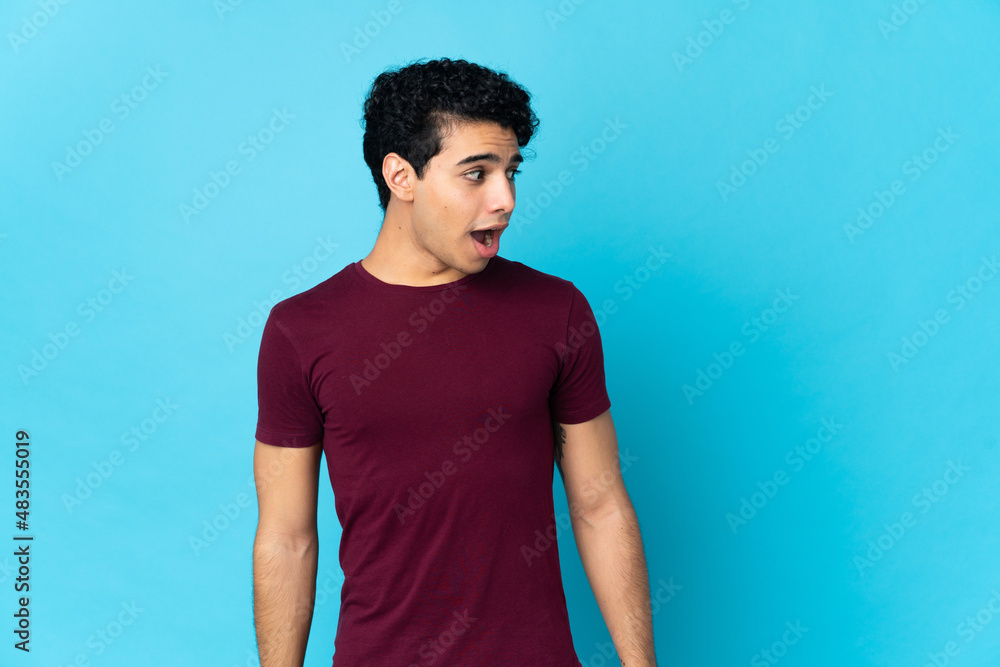 Young Venezuelan man isolated on blue background doing surprise gesture while looking to the side Young Venezuelan man isolated on blue background doing surprise gesture while looking to the side
