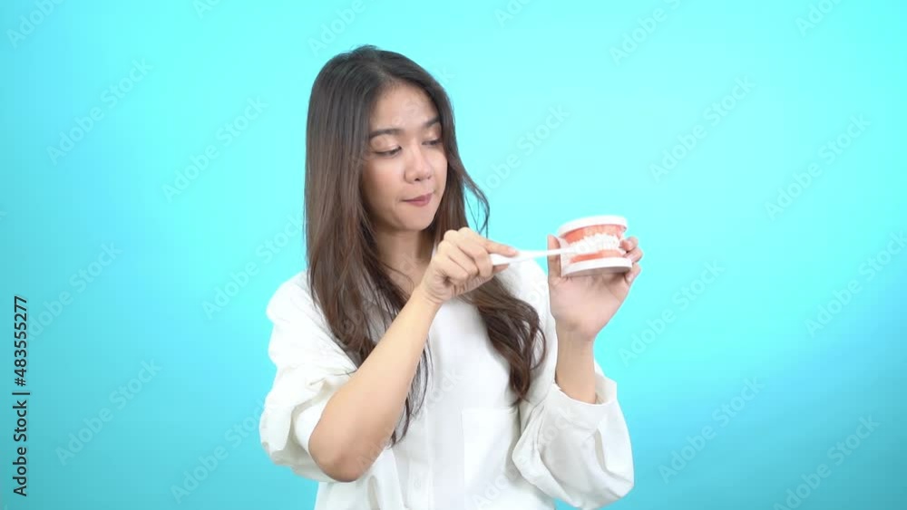 Beautiful Asian woman holding a toothbrush and a dental teeth model showing how to brush properly both upper and lower front teeth at camera with a happy smile on face isolated on turquoise background Beautiful Asian woman holding a toothbrush and a dental teeth model showing how to brush properly both upper and lower front teeth at camera with a happy smile on face isolated on turquoise background