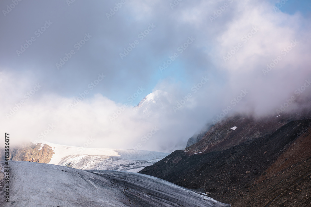 Scenic mountain landscape with large glacier in low clouds in sunrise colors. Colorful view to glacier tongue in morning sunlight in low clouds. Atmospheric high mountain scenery at early morning.