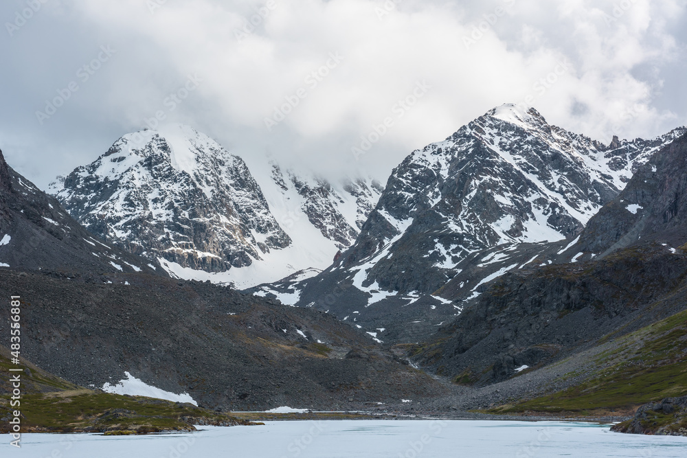 Dramatic mountain landscape with frozen alpine lake and high snow mountains and sunlit big rocks among low clouds. Atmospheric scenery with ice lake on background of large snowy mountains in overcast.