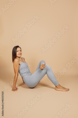 Wallpaper Mural Young woman is doing yoga. Side view slender brunette in gray top and leggings smiles and stands with her hands on floor and one leg on her knee, she poses on neutral beige background. Torontodigital.ca