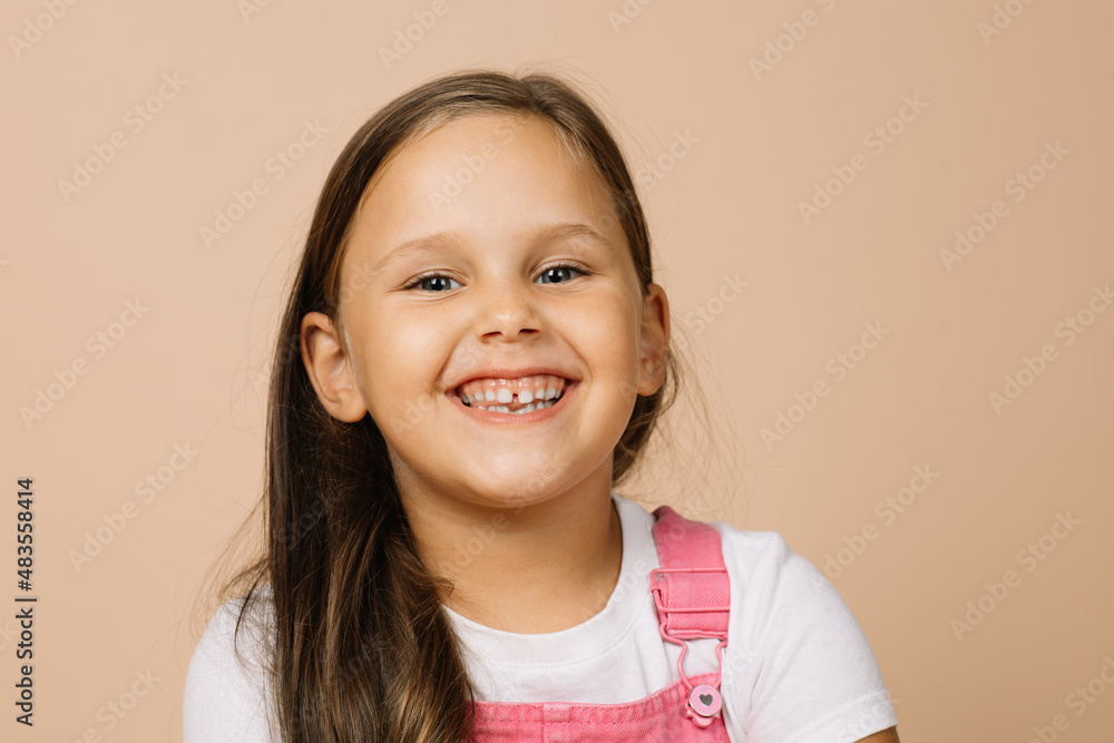 Little girl with bright shining eyes and excited blissful smile with teeth looking at camera wearing bright, pink jumpsuit and white t-shirt on beige background. Little girl with bright shining eyes and excited blissful smile with teeth looking at camera wearing bright, pink jumpsuit and white t-shirt on beige background.