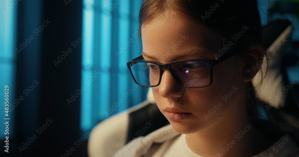 Face of serious smart teenage girl wearing glasses to protect her eyes while working on computer. Young college student is focused on studying online from home late at night. Close up shot. Face of serious smart teenage girl wearing glasses to protect her eyes while working on computer. Young college student is focused on studying online from home late at night. Close up shot.