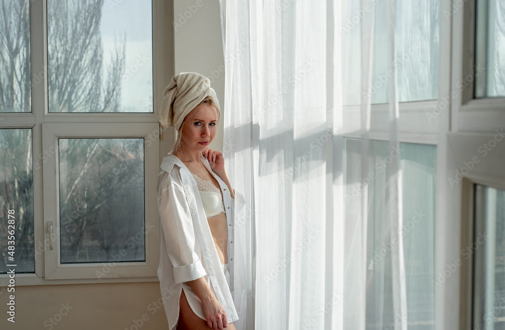 a girl in underwear and a white shirt with a towel on her head stands near the window