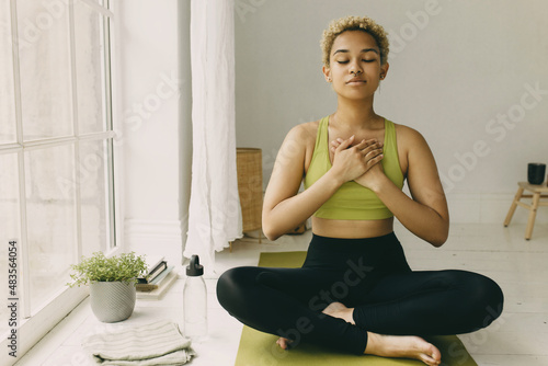 Fototapeta Naklejka Na Ścianę i Meble -  Young African American lady practicing yoga at home in black leggings sitting on floor in lotus posture hands crossed on chest slight delight smile on face, feeling her body. Emotional health