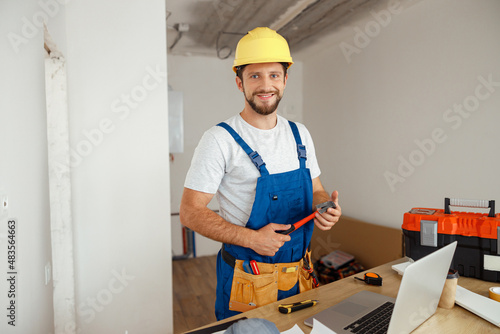 Professional handyman worker in uniform wearing tool belt and hardhat smiling at camera, holding hammer during renovation work indoors