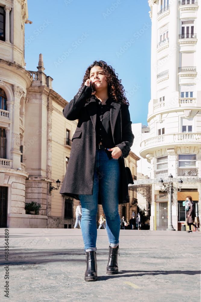 Naklejka premium Smiling elegant young pretty woman talking on the phone while walking in the city on a sunny day