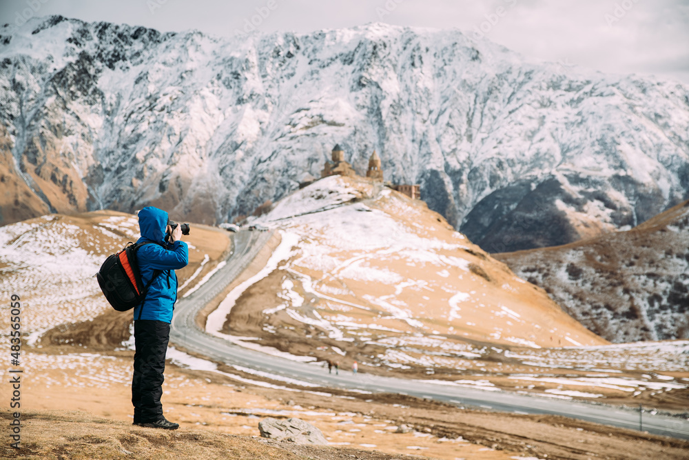 Stepantsminda, Gergeti, Georgia. Man Tourist Backpacker Traveler Photographer Taking Pictures Photos Of Holy Trinity Church - Tsminda Sameba. Beautiful Georgian Landscape In Early Winter.