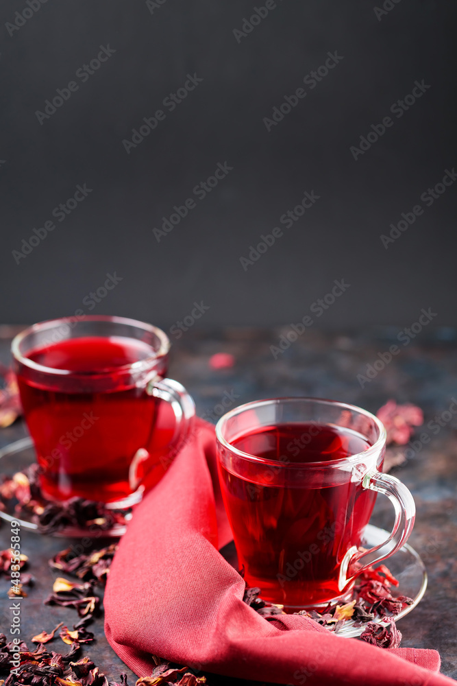 Hibiscus tea in glass cup and burgundy napkin. Cup of red hibiscus tea and dry hibiscus petals on dark background. Healthy slimming drink. Copy space Hibiscus tea in glass cup and burgundy napkin. Cup of red hibiscus tea and dry hibiscus petals on dark background. Healthy slimming drink. Copy space